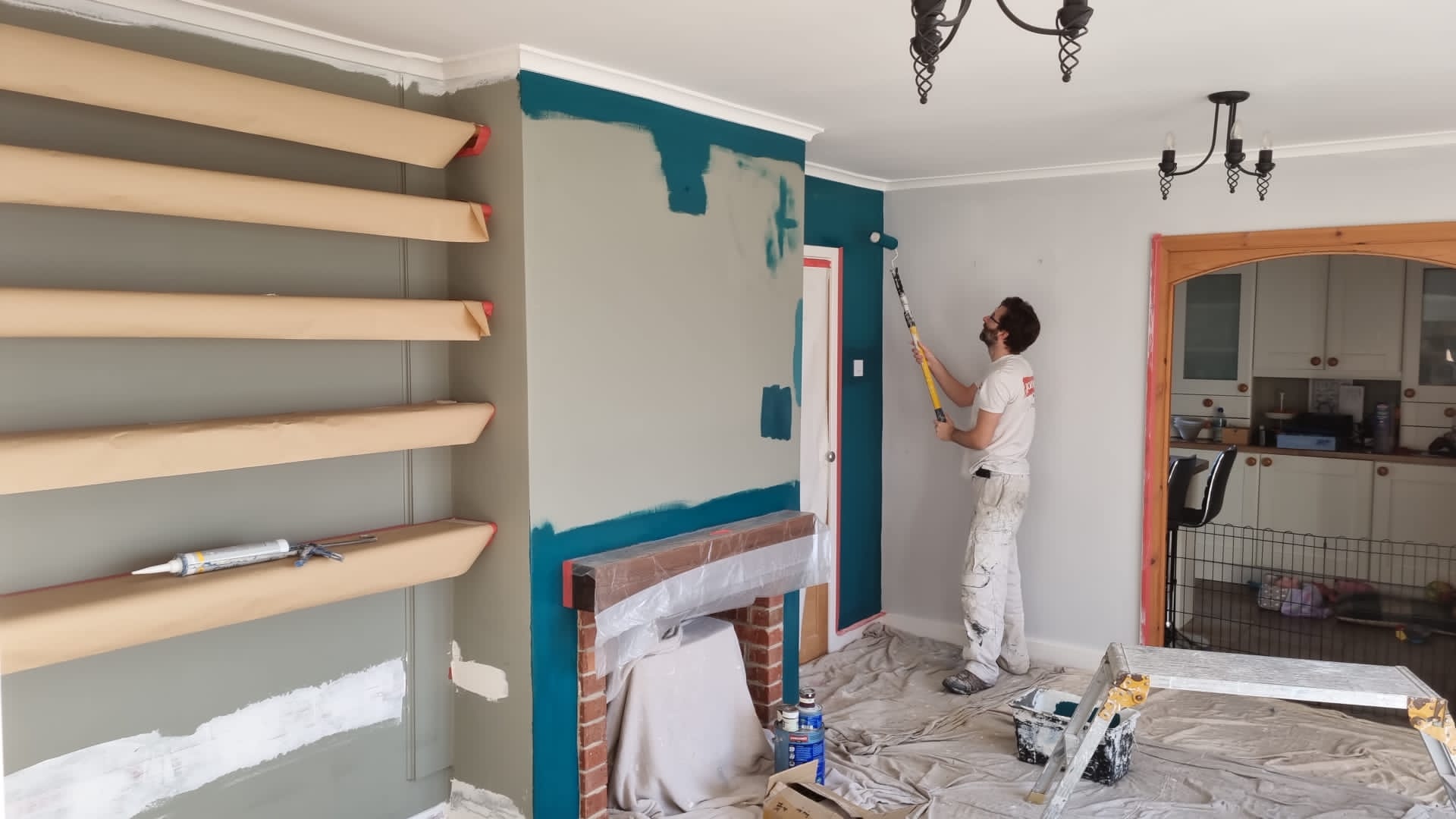Decorator in whites rolling paint onto a wall, with dust sheets down and shelving masked off